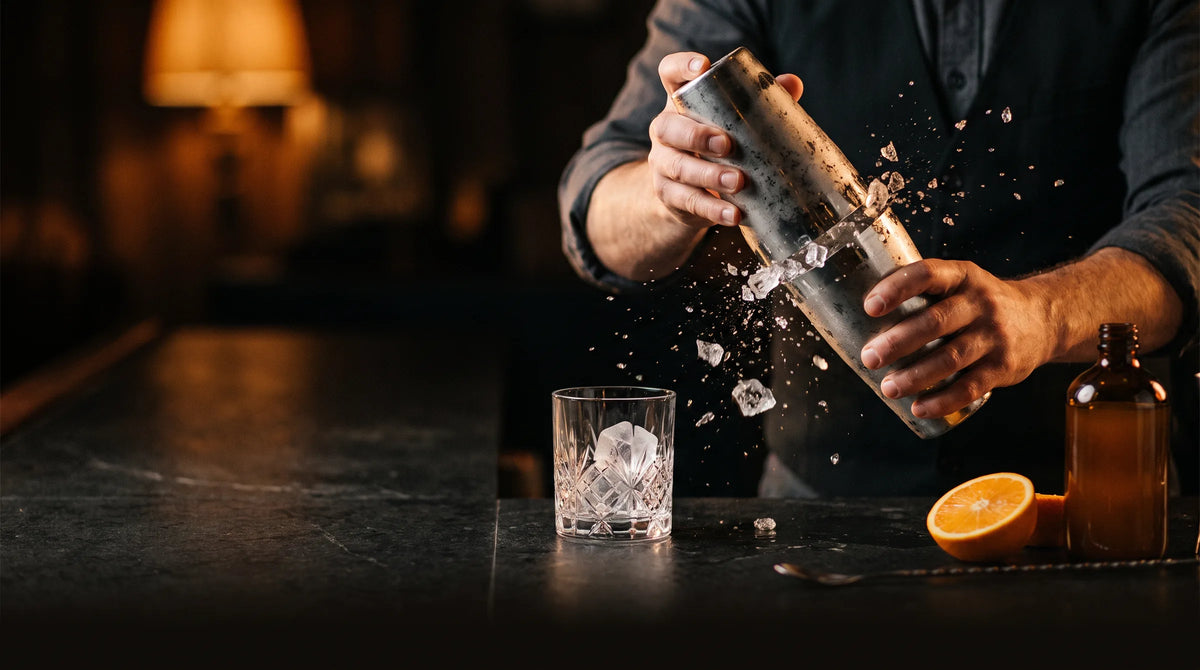 A bartender shaking a cocktail shaker over a rocks glass with ice and an orange slice