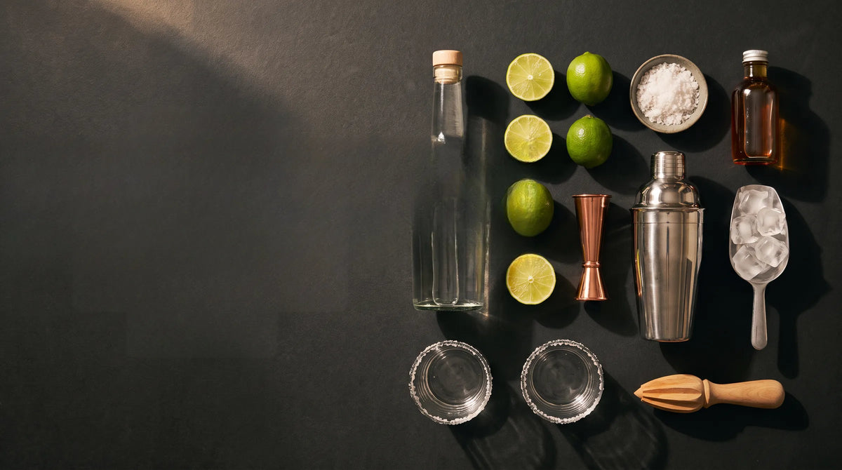 Margarita ingredients laid out on slate: a bottle of tequila, limes, a shaker, a jigger and a citrus press