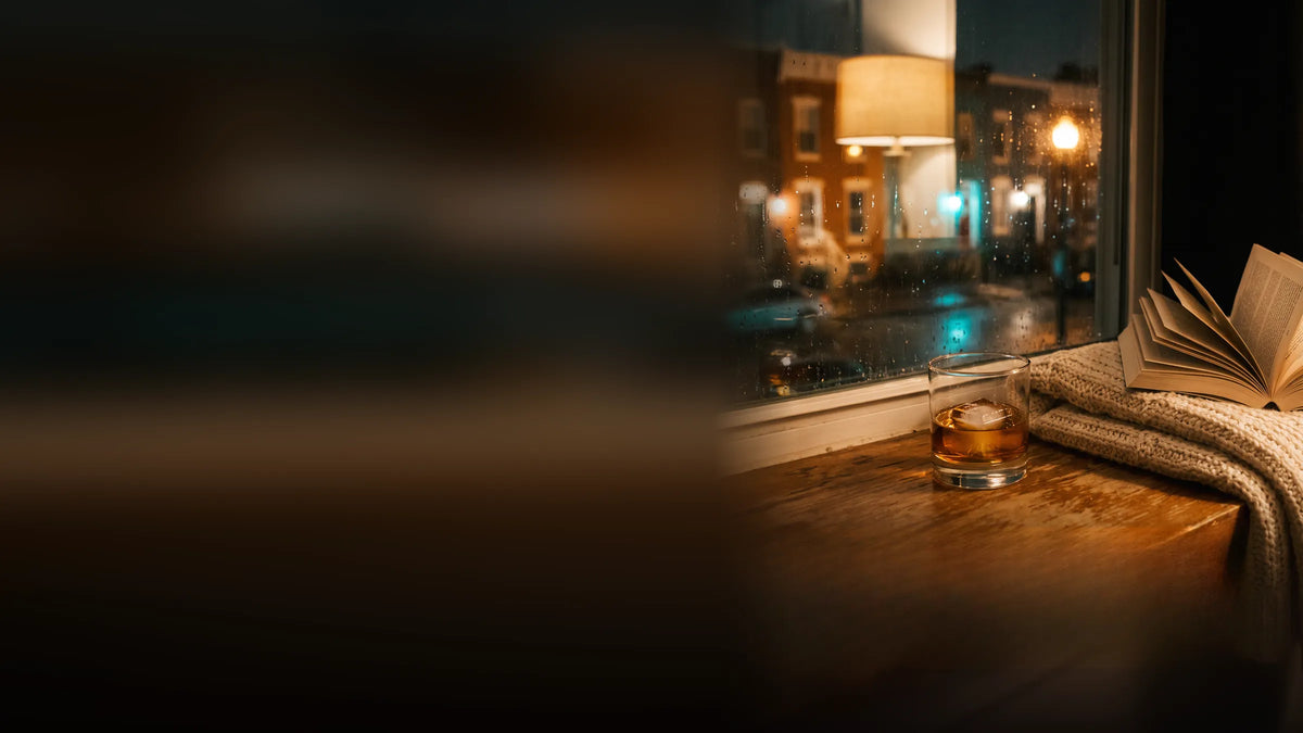 A glass of whisky on a window ledge beside a book and a blanket, with rain on the window